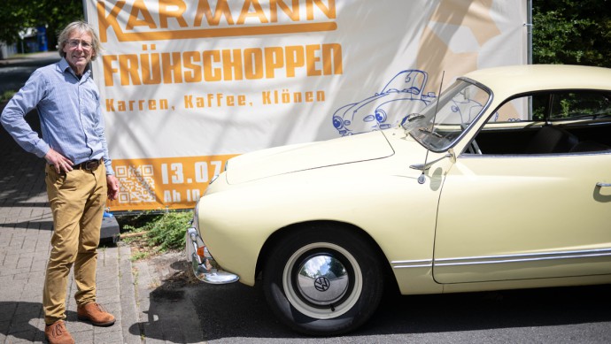 Joachim Acker, Vorsitzender vom Verein Karmannstadt, steht vor einem Plakat, das ein Karmann-Treffen ankündigt.