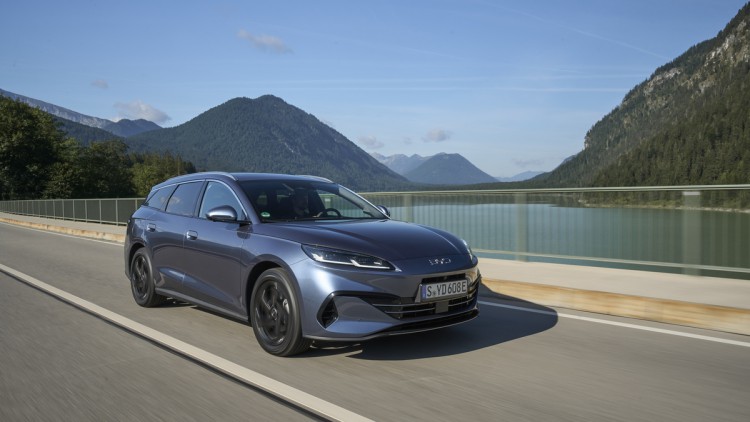 BYD Seal 6 DM-i Touring fahrend schräg von vorn auf einer Brücke über einem See in den Voralpen