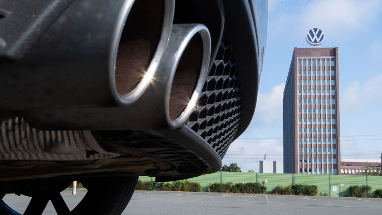 Auspuffrohre eines Volkswagen, fotografiert mit dem Markenhochhaus am VW-Werk Wolfsburg im Hintergrund. 