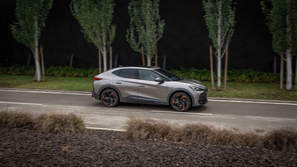 Cupra Tavascan VZ fahrend von der Seite fotografiert vor Bäumen und einer Fabrikwand